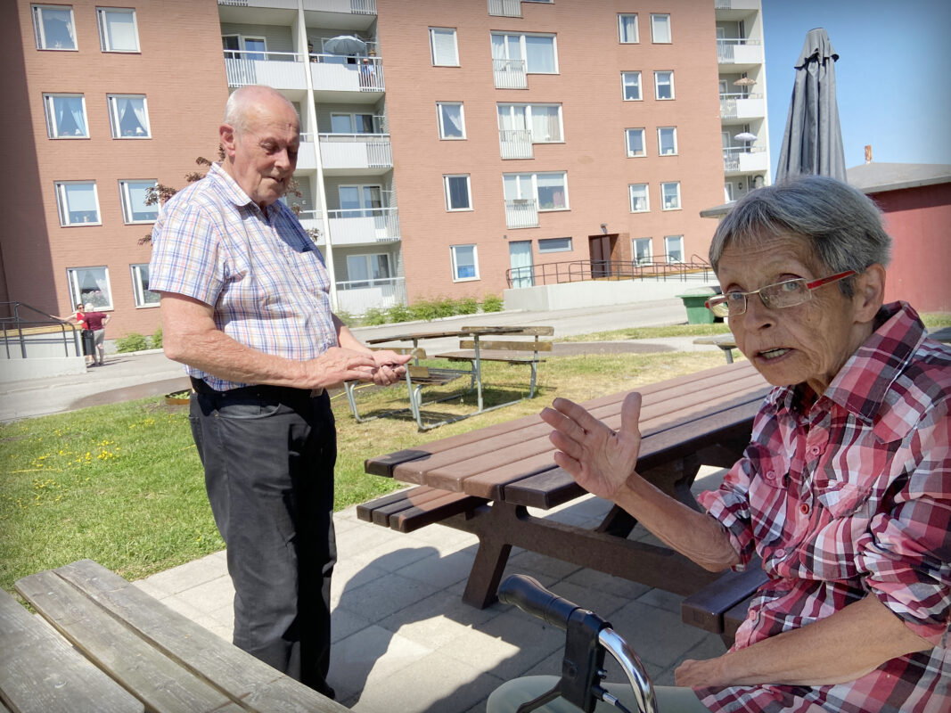 En äldre kvinna i förgrunden och en äldre man i bakgrunden, utomhus i ett höghusområde.