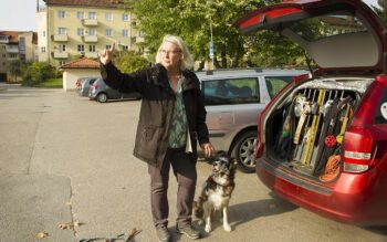 Foto på Marie Larsson, med en av sina hundar (Australian shepard) bredvid sig, och en som sticker ut nosen genom gallret i den öppna bakluckan på en röd bil. Marie har axellångt ljust/vitt hår, glasögon, grönmönstrad topp, svart jacka och svarta byxor. Hon gestikulerar åt vänster i bild med ett finger. i bakgrunden syns parkeringen och ett ljusgult fyravåningshus med putsad fasad.