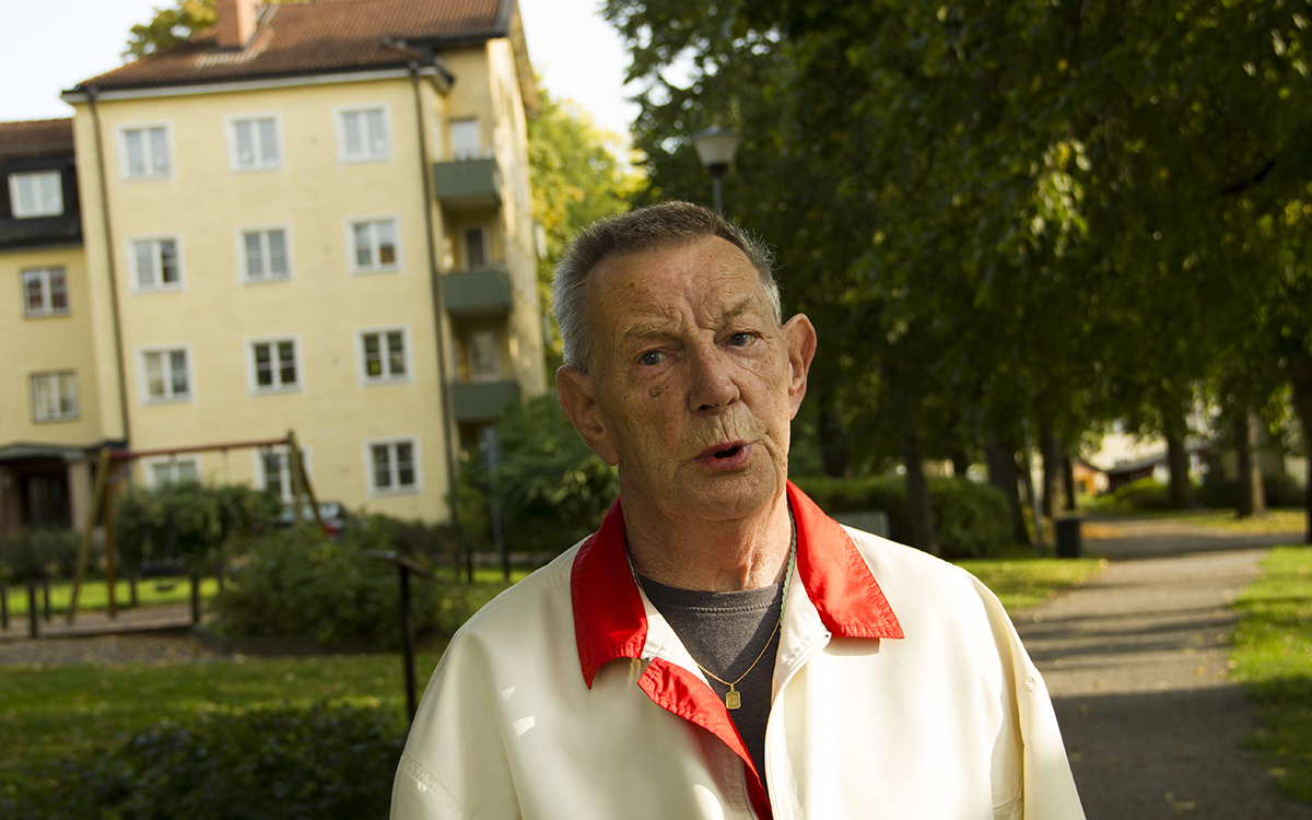 Halvkroppsporträtt på Ulf Boholm. Han har kortklippt grått hår och en ljus jacka med röd krage. I bakgrunden syns träd och annan grönska och ett gult putsat fyravåningshus med gröna balkonger.