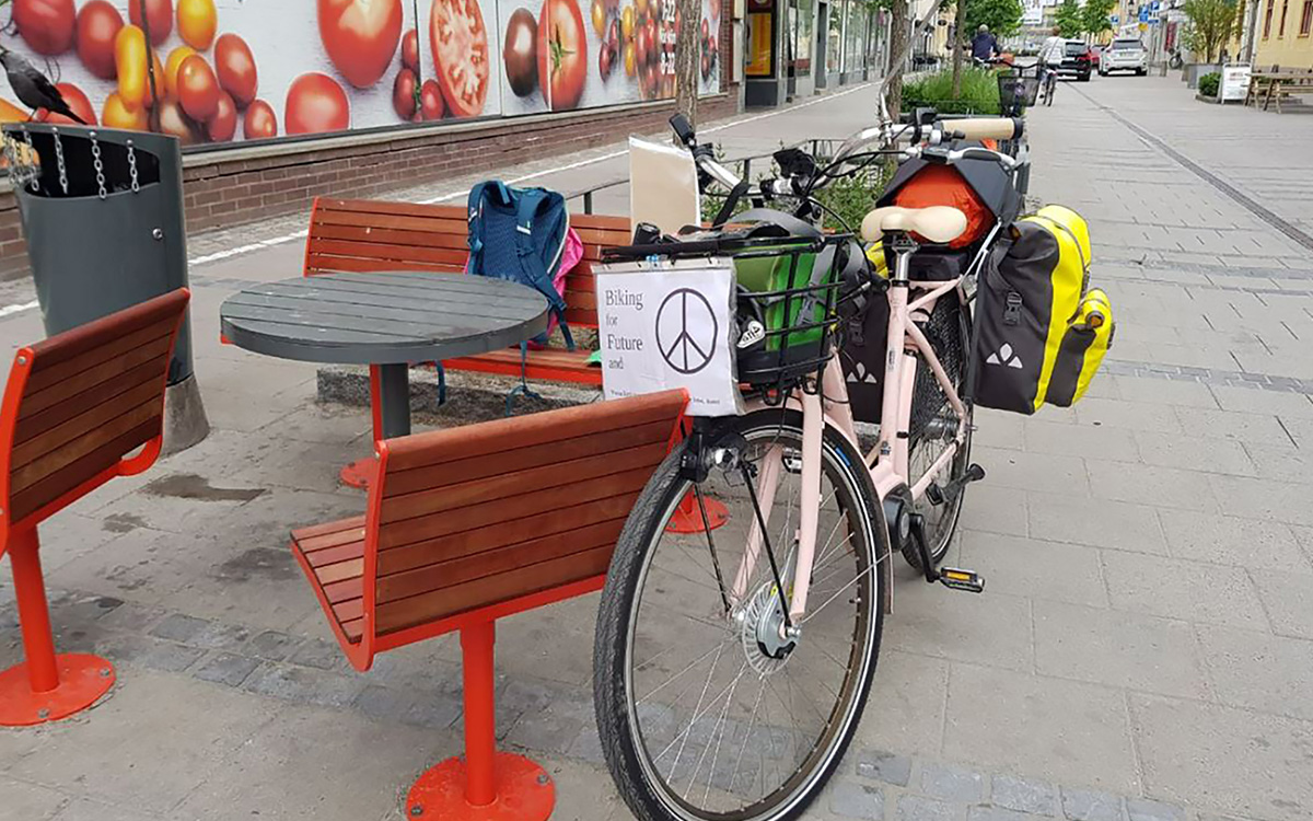 Dorothees cykel med packväskorna lutad mot en stol och en bänk på en gata i Sverige. På cykelkorgen ser man en lapp där det står Biking for future and ... följt av fredsmärket.