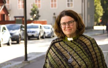 Halvkroppsporträtt på Giselle Hedman som har axellångt rödbrunt hår, glasögon, grön skjorta med vit "prästkrage" under en stickad poncho/sjal i gröna toner. I bakgrunden, suddigt, en gata med parkerade bilar, en ljus husfasad som ser ut att vara mer än hundra år gammal och grönska till höger bortom huset. Giselle Hedman arbetar vräkningsförebyggande som diakon och har hjälpt åtskilliga hyresgäster som riskerat vräkning.