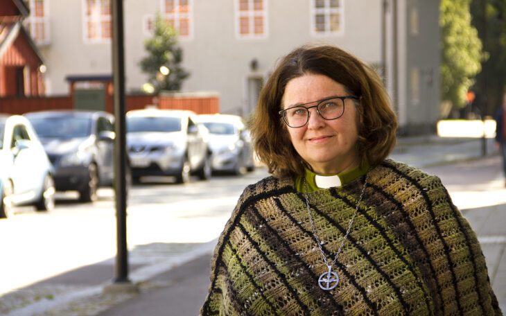 Halvkroppsporträtt på Giselle Hedman som har axellångt rödbrunt hår, glasögon, grön skjorta med vit "prästkrage" under en stickad poncho/sjal i gröna toner. I bakgrunden, suddigt, en gata med parkerade bilar, en ljus husfasad som ser ut att vara mer än hundra år gammal och grönska till höger bortom huset. Giselle Hedman arbetar vräkningsförebyggande som diakon och har hjälpt åtskilliga hyresgäster som riskerat vräkning.