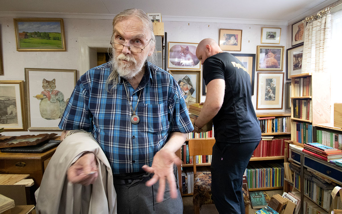 Kenneth Jälkhammar står i sitt bibliotek medan Charlie Knutsson från Flyttprinzen plockar böcker i bakgrunden.