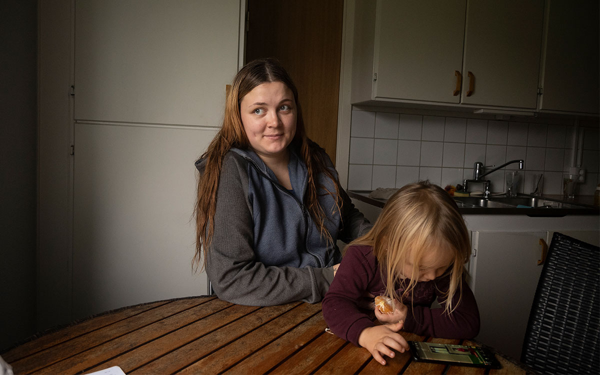 En ung kvinna med ett barn i knät vid ett köksbord i en lägenhet. Barnet äter en citrusfrukt och tittar på en mobiltelefon.