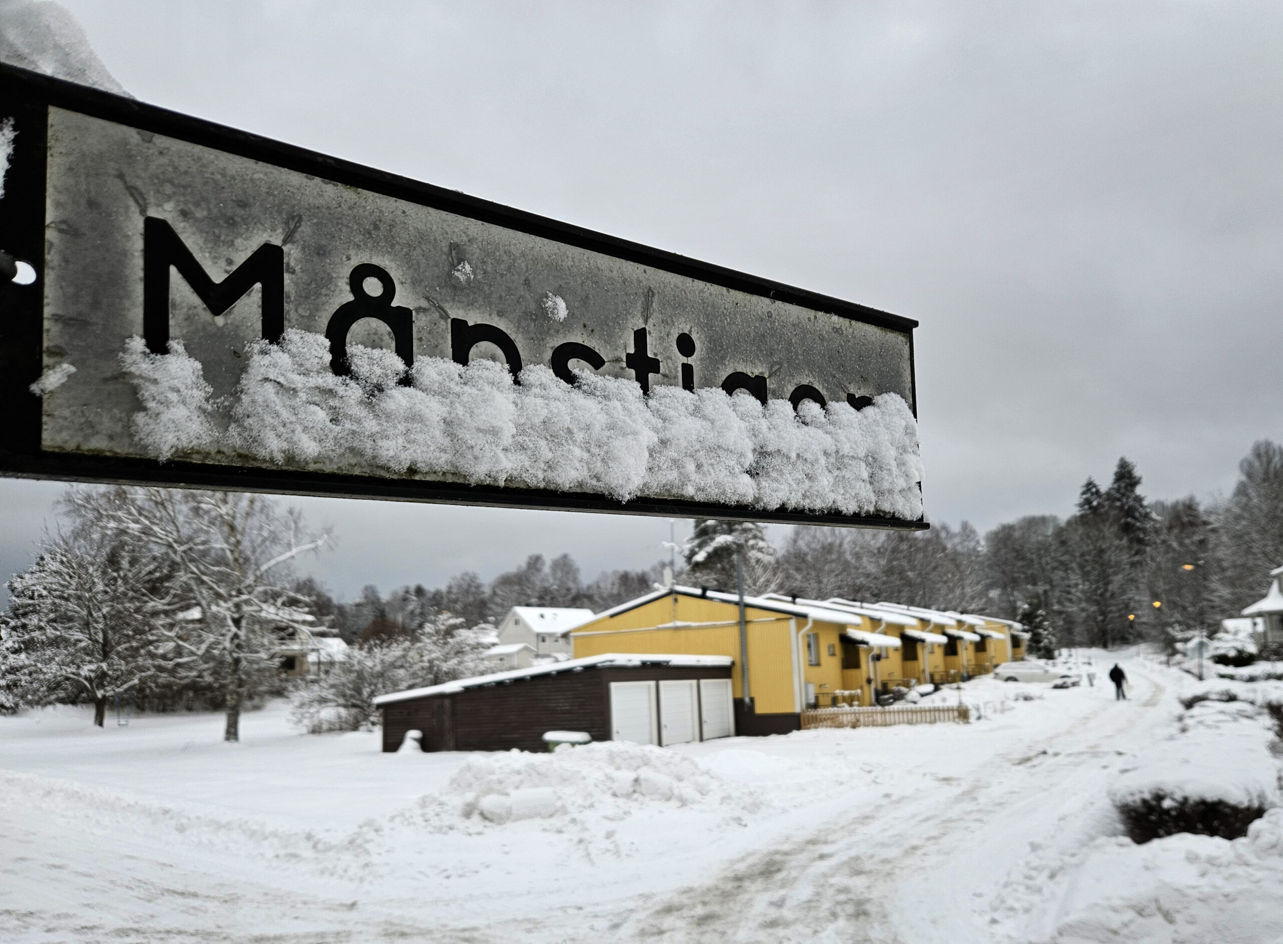 En vit och svart skylt med texten "Månstigen" som döljs av snö i nederkant. I bakgrunden några gula hyresradhus.