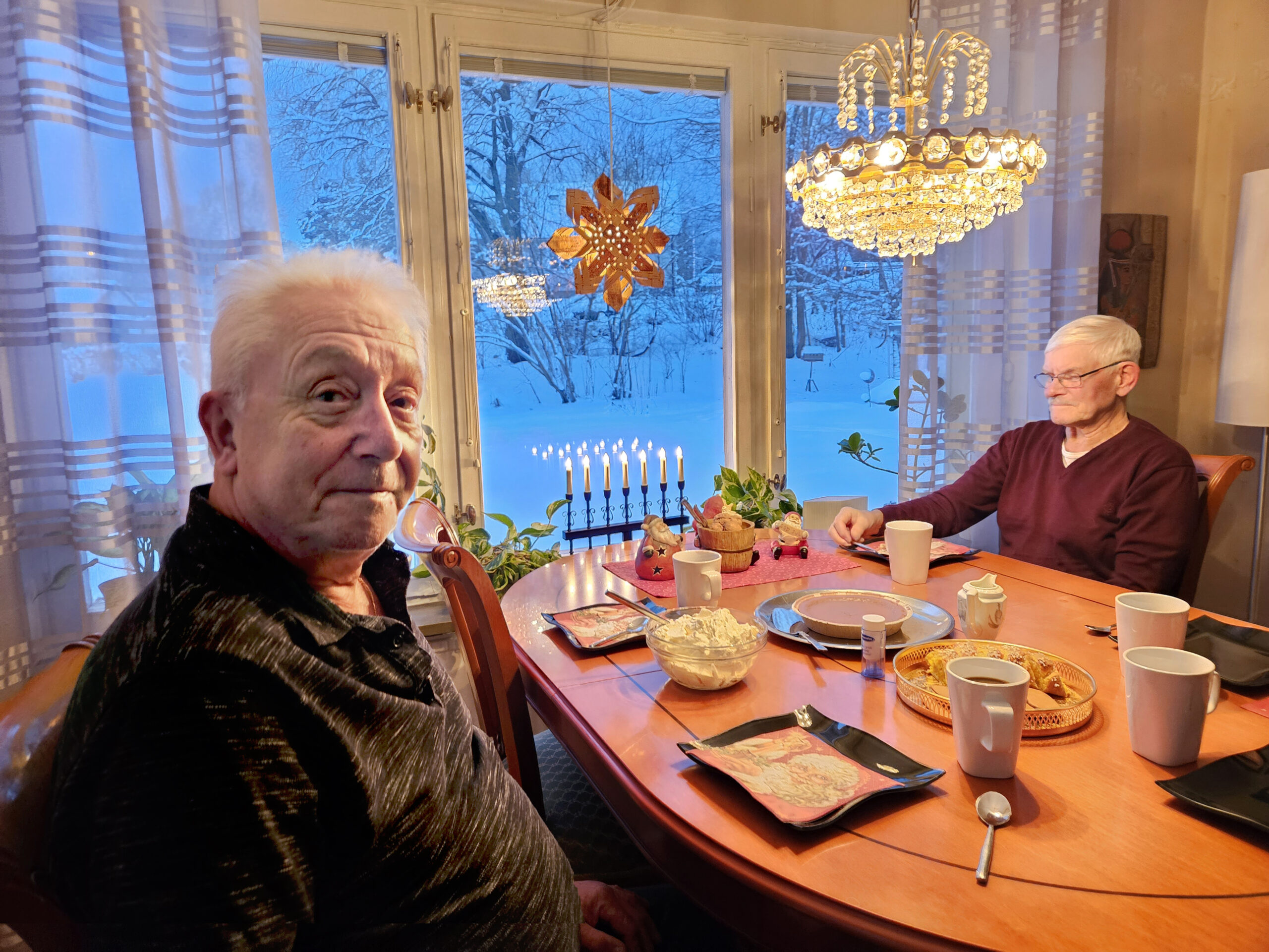 Två män sitter vid ett adventsfikadukat bord. I fönstret hänger en julstjärna och utanför syns snöklädda buskar.