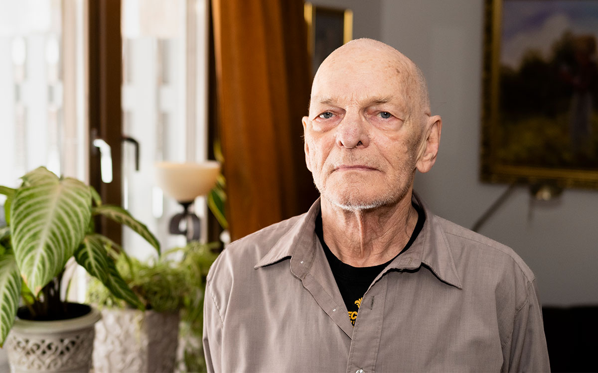 En skallig äldre man med grå skjorta som står i ett vardagsrum.