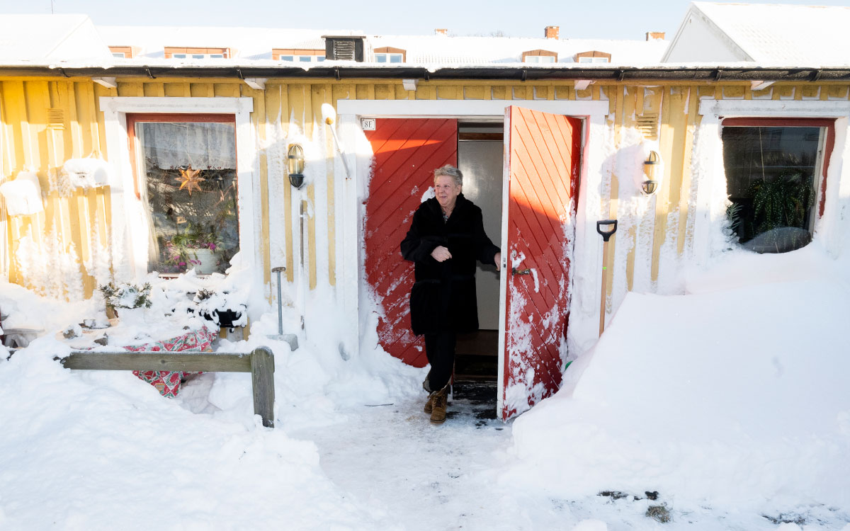 En äldre, kortklippt kvinna som går ut genom en röd port till ett gult hus med snöig fasad.