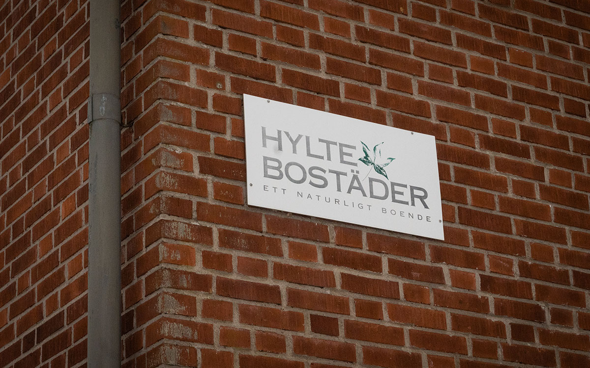 En skylt med texten "Hyltebostäder ett naturligt boende" på ett hus med tegelvägg.