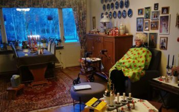 En äldre dam som sitter i sitt vardagsrum i en fåtölj och under en filt. På väggarna flera fotografier av barn samt prydnadstallrikar. Utanför fönstret ett snöigt landskap och en julljusstake i fönstret.