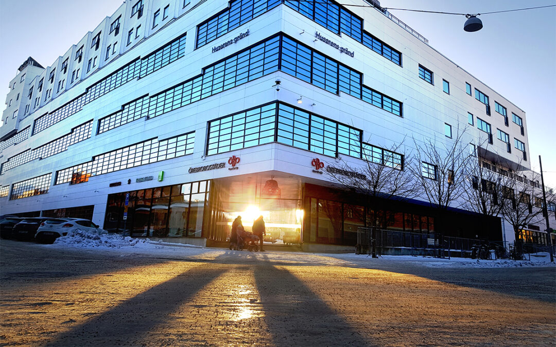 Personer i starkt motljus vid Örebrobostäders vita fasad. Vinterväg och skuggor över gatan.