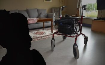 På bilden ser man en rollator som står mitt på golvet i ett rum. I bakgrunden syns bland annat en soffa och ett fönster. En svart silhuett föreställande en kvinna är inflikad i bilden.