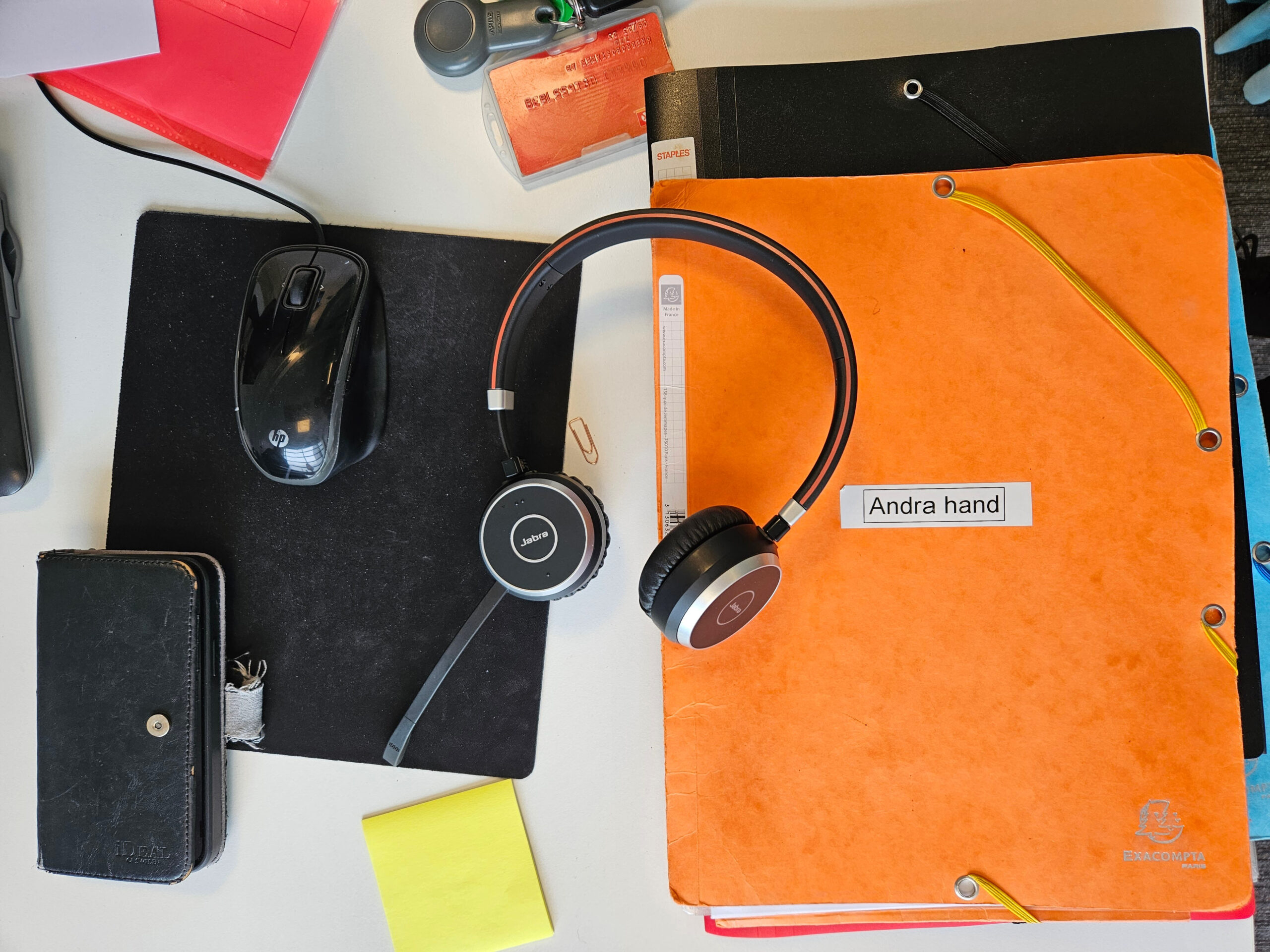 Detaljbild av saker på ett skrivbord: mappar, gem, post-it-lapp, mobil, hörlurar och inpaseringstagg.