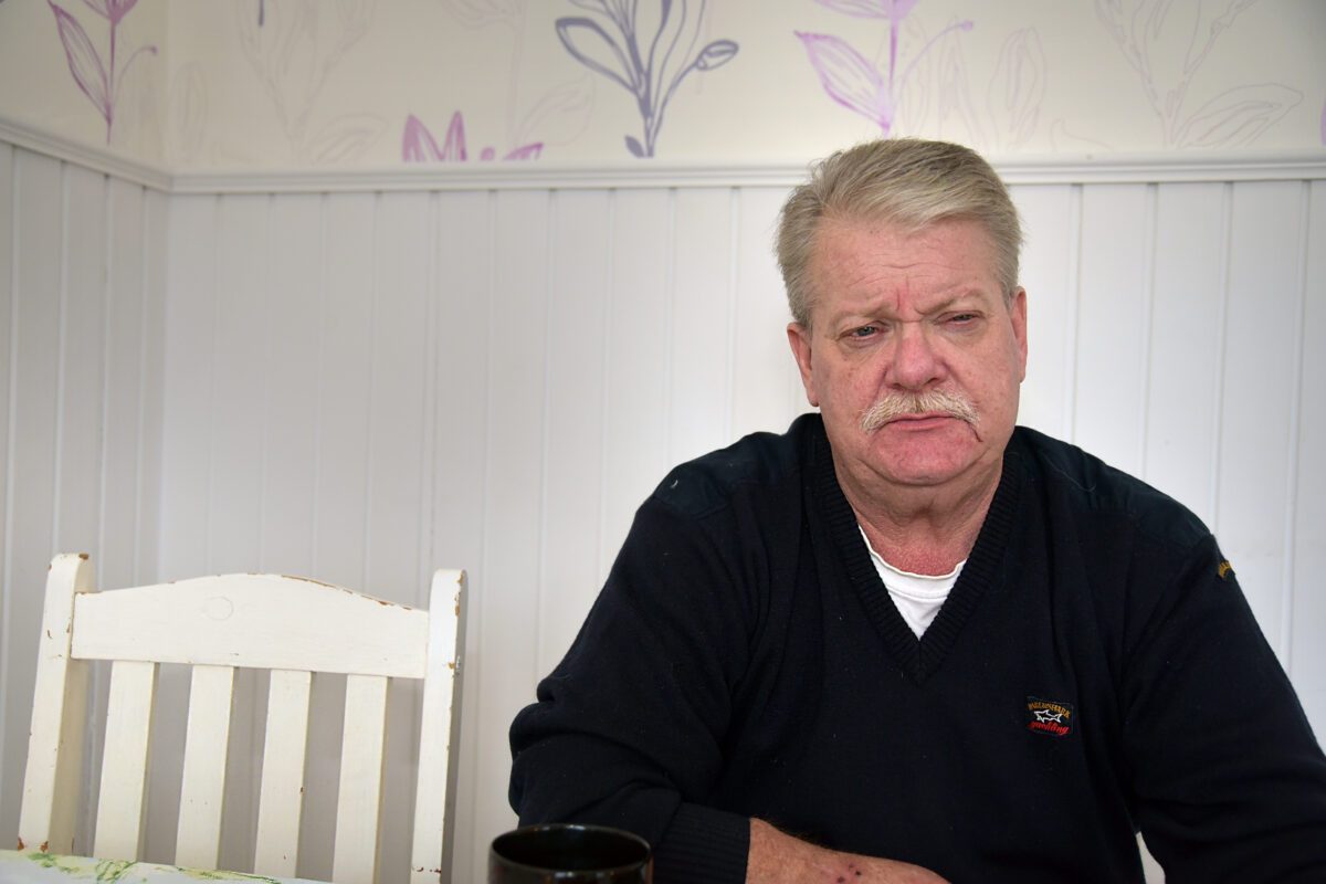 En man i 60-årsåldern sitter vid ett bord med en vitmålad trästol bredvid sig.