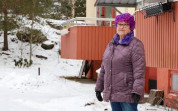 Kvinna som står utanför ett hyreshus. En röd balkong syns i bakgrunden. Det är snö på marken.