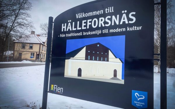 Bilden visar en välkommen till Hälleforsnäs-skylt som går mestadels i blått. På skylten syns en bruksbyggnad.