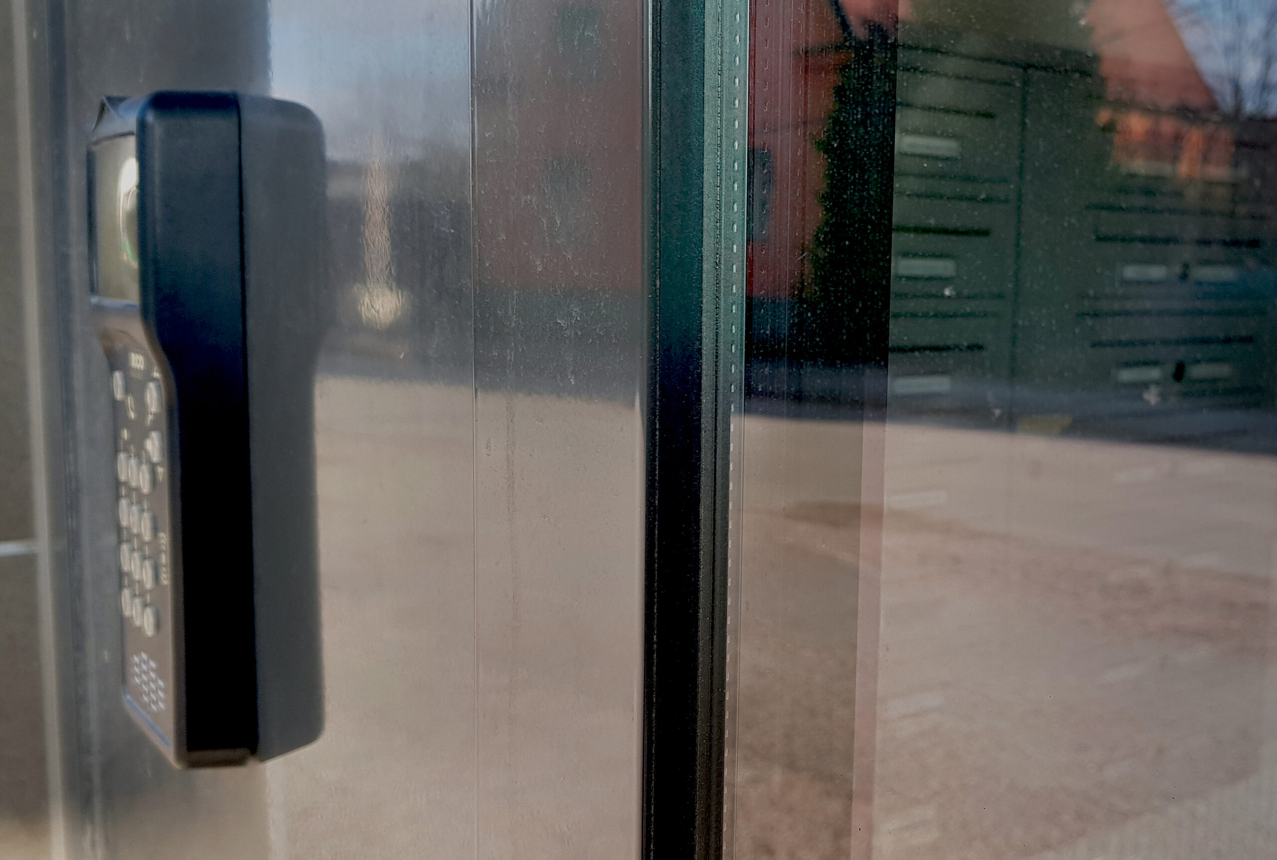 Ett foto genom en ruta där ett hus reflekteras och en taggläsare med kod syns i förgrunden. Innan för rutan syns postlådorna i ett lägenhetshus.