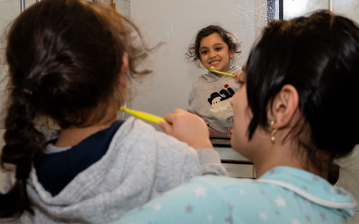 En ung kvinna som håller runt en liten pojke som borstar tänderna och ser på sig själv i en badrumsspegel.
