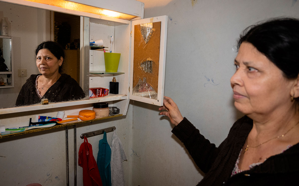 En äldre kvinna som öppnar ett badrumsskåp som är slitet och där en spegel är trasig.