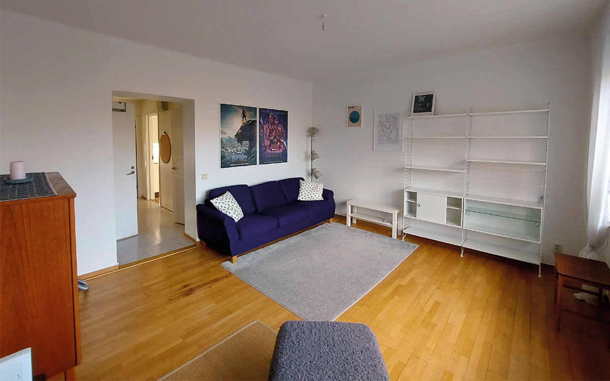 Ett vardagsrum i en möblerad lägenhet med soffa, skåp, matta, tavlor på väggarna och en stringhylla.