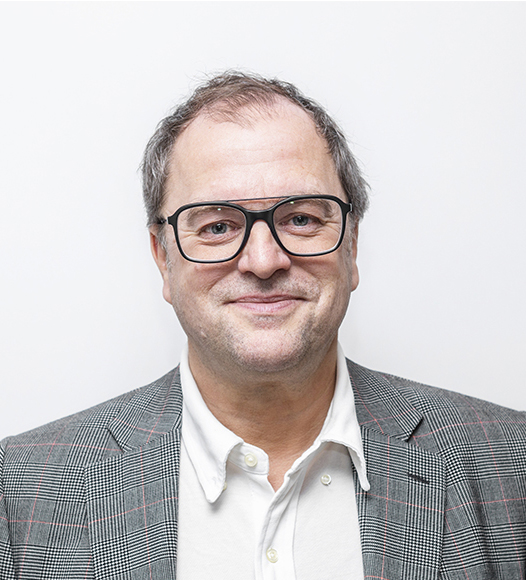 Porträtt av en man som ler mot kameran. Han har svarta glasögon, mörkt tunt hår och bär vit skjorta och grå jacka.