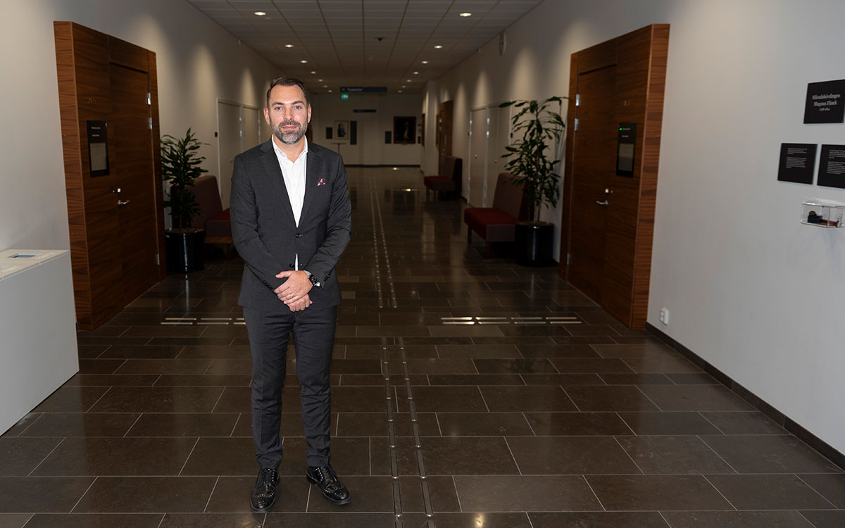 En man i skägg, kostym och vit skjorta som står med händerna knäppta i en korridor i en domstolsbyggnad.