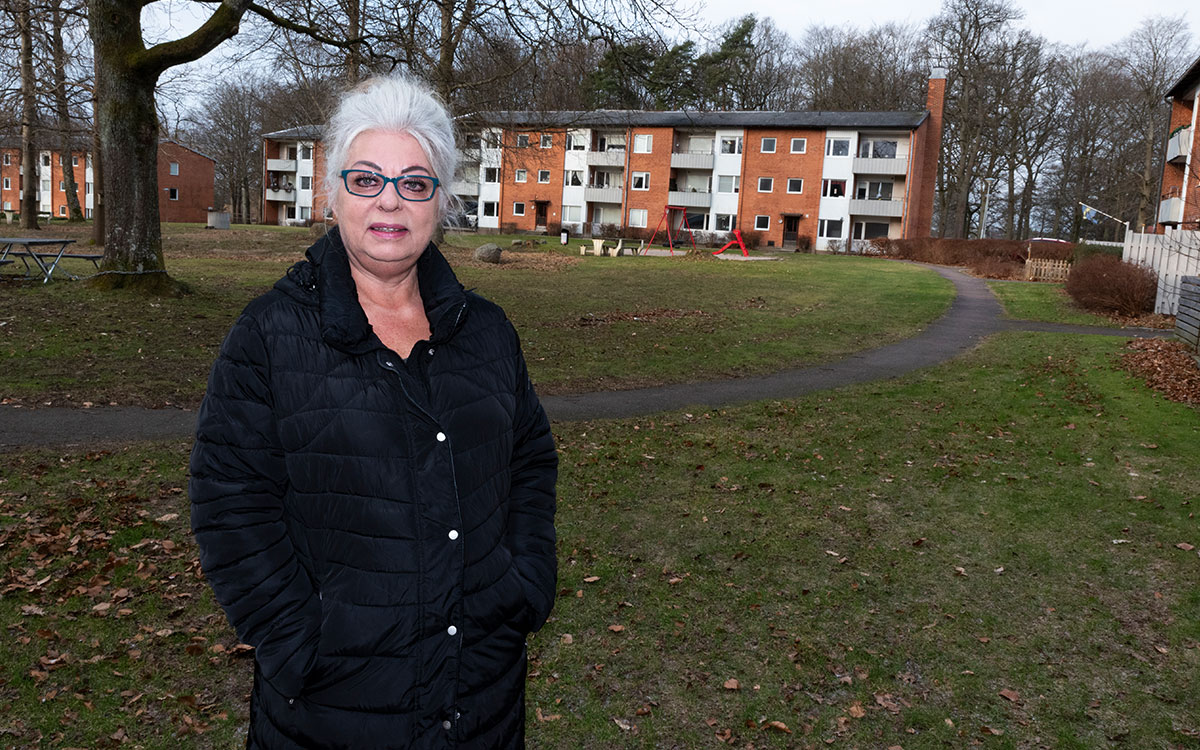 En äldre kvinna med glasögon och vitt hår som står framför ett trevåningshus.