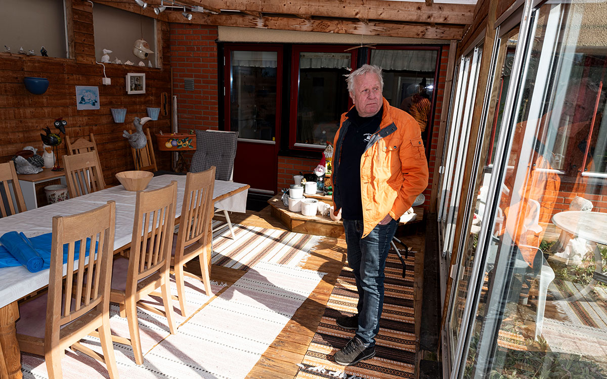 En äldre man i en orange jacka som står i ett uterum och speglas i glasdörren.