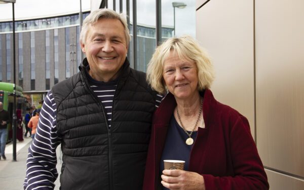 Halvkroppsporträtt på Jan Rydenäs och Annika Backlund som står intill en kopparfärgad fasad med fönster. I bakgrunden en grå böjd fasad, en grön buss och människor i rörelse på trottoaren. AB-hem har vänt sig till hyresnämnden för att få höja hyran i Fjärdhundra.