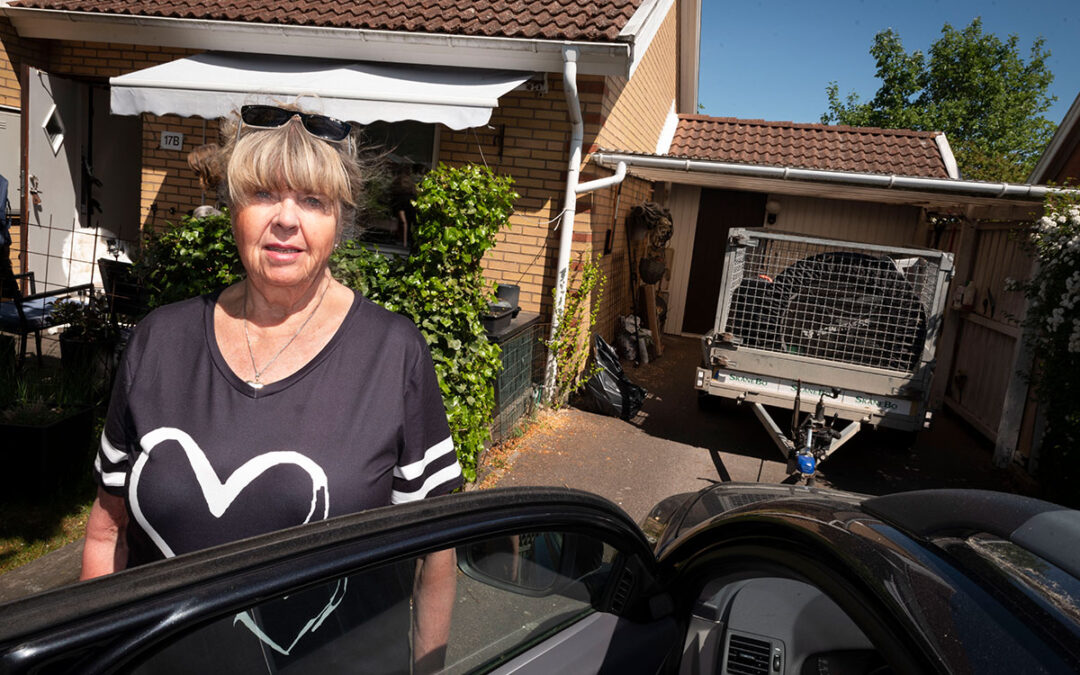 En äldre kvinna som står vid en bil med öppen dörr framför en carport med ett släp i.