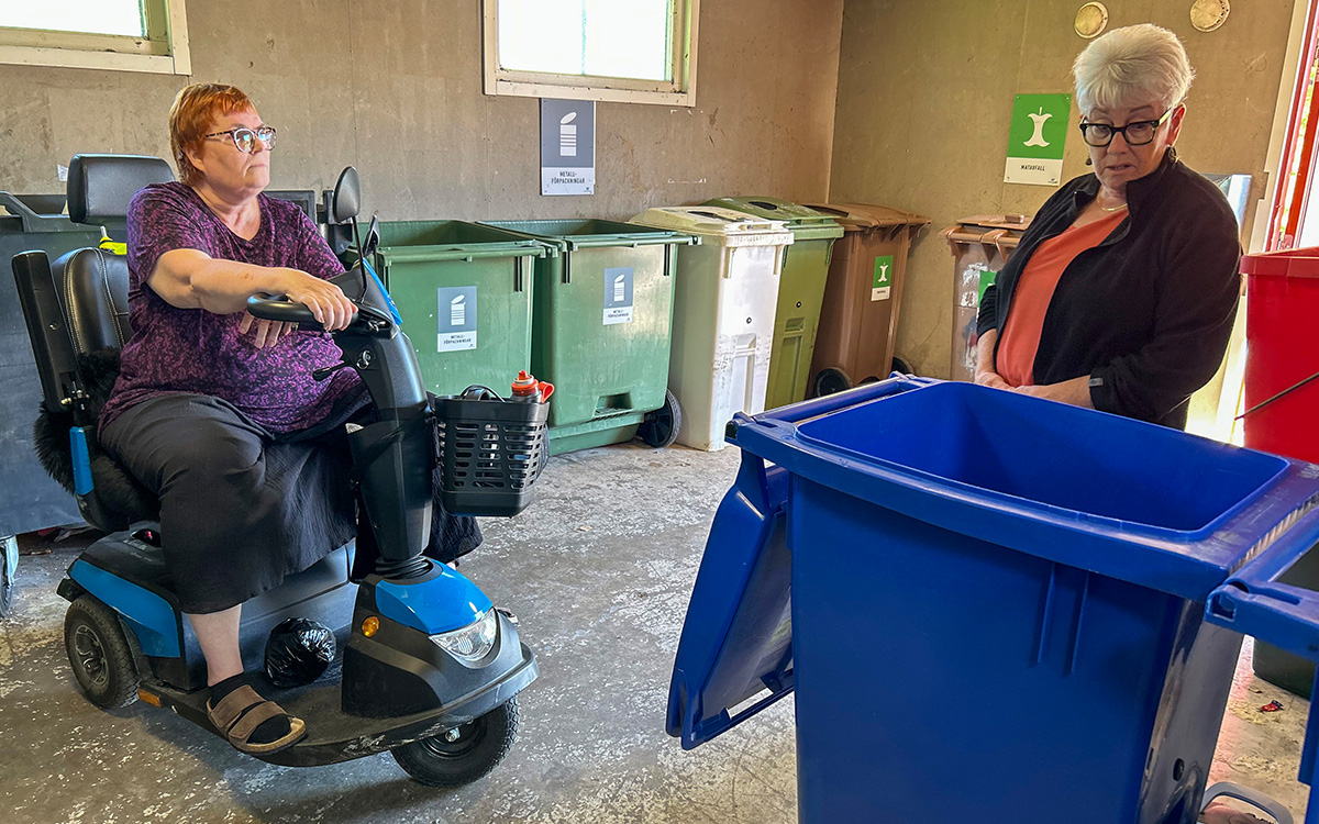 Bilden visar två kvinnor i ett miljörum med flera containrar och behållare En av kvinnorna sitter i en elrullstol.