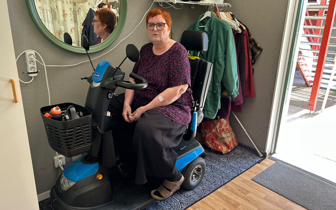 Kvinna i en elrullstol. Hon sitter i hallen, bredvid en spegel. En del av hennes ansikte syns i spegeln. Ytterdörren står öppen.