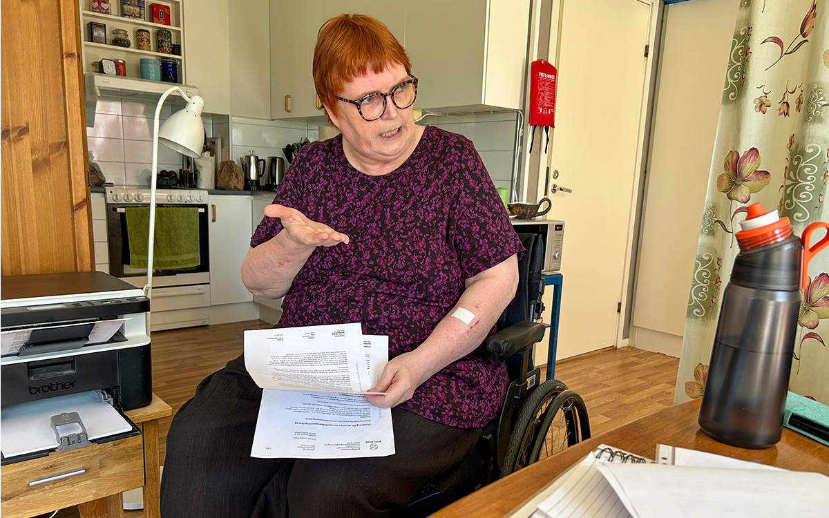 Kvinna sim sitter i en rullstol. Hon gestikulerar med ena handen, i den andra håller hon handlingar från kommunen.