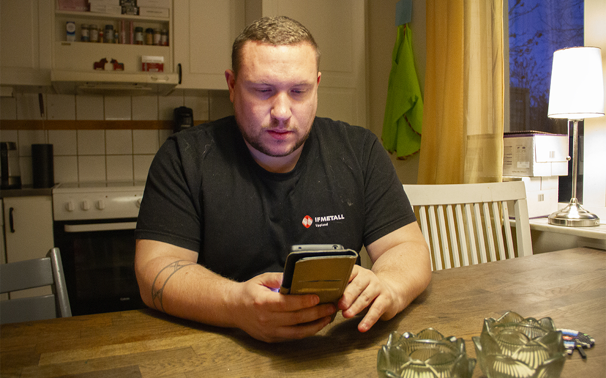 Jacop Lindholm sitter vid ett köksbord med armarna på borde och tittar ner i mobilen han håller i. Svart t-shirt med en liten logga: IF Metall. Till höger i fönstret en bordslampa. AB-hem vill höja hyrorna i Fjärdhundra genom en bruksvärdering.