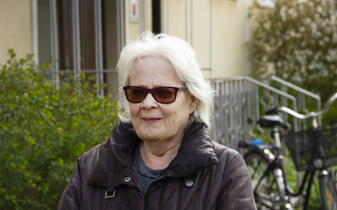 Halvkroppsporträtt på Annika Hymark som har vitt axellångt hår, bruna solglasögon och brun jacka. I bakgeunden syns en ljust snadgul putsad fasad, ett räcke i metall, en cykel och buskar. Uppsalahem vill riva Annika Hymarks hus i kvarteret Idun i Svartbäcken i Uppsala.