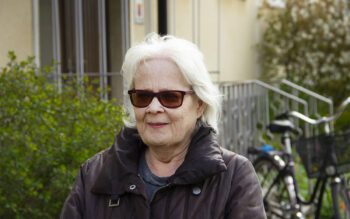 Halvkroppsporträtt på Annika Hymark som har vitt axellångt hår, bruna solglasögon och brun jacka. I bakgeunden syns en ljust snadgul putsad fasad, ett räcke i metall, en cykel och buskar. Uppsalahem vill riva Annika Hymarks hus i kvarteret Idun i Svartbäcken i Uppsala.
