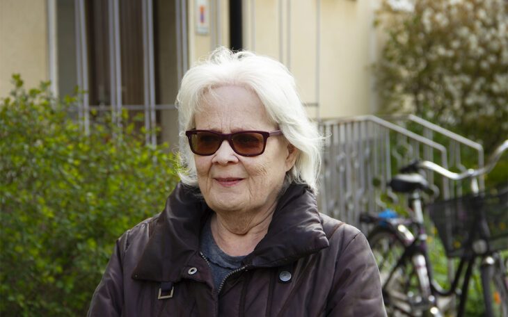 Halvkroppsporträtt på Annika Hymark som har vitt axellångt hår, bruna solglasögon och brun jacka. I bakgeunden syns en ljust snadgul putsad fasad, ett räcke i metall, en cykel och buskar. Uppsalahem vill riva Annika Hymarks hus i kvarteret Idun i Svartbäcken i Uppsala.