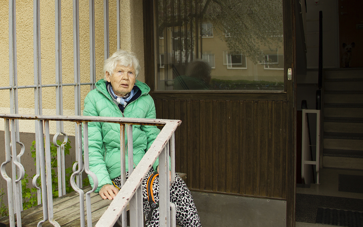Maj Landsberg sitter på bänken vid ingången till sin trappuppgång. Hon har gråvitthår i en kort frisyr, en grönturkos täckjacka och svart- och vitspräckliga byxor. I förgrunden ett järnräcke. Till vänster syns den sandfärgade putsade fasaden och huset mitt emot av samma typ och ett träd speglas i ett glasparti intill porten. Maj landsberg bor i ett av de hus i kvarteret Idun i Svartbäcken som Uppsalahem vill behålla och renovera.