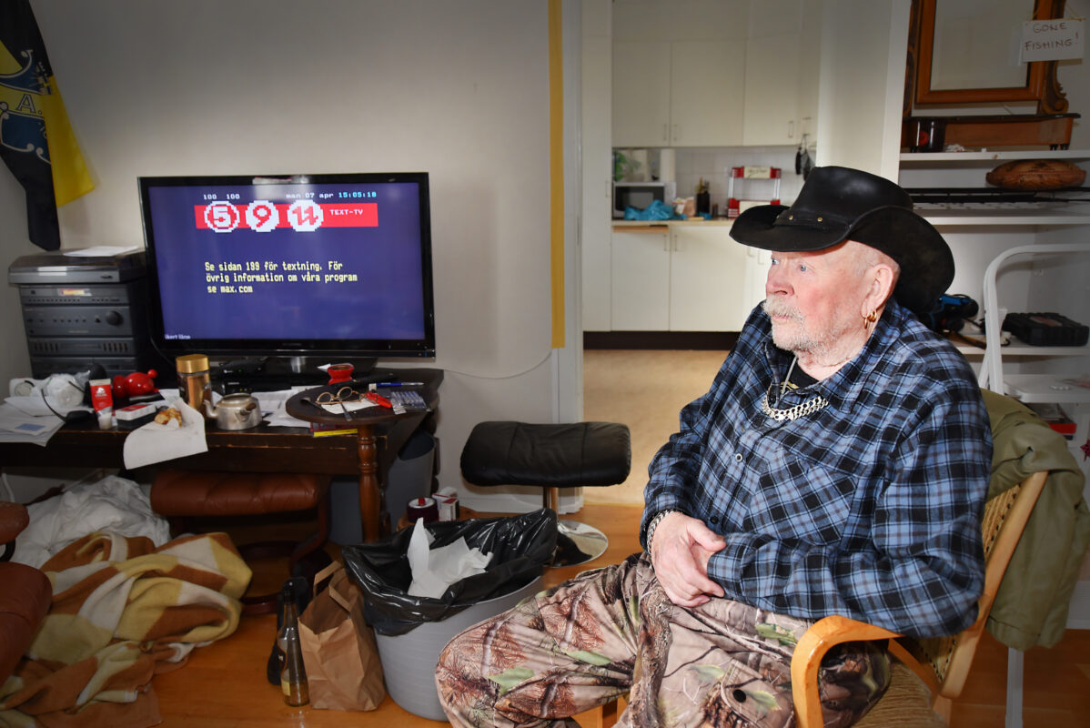 En äldre man med cowboyhatt i en fåtölj i ett vardagsrum med tv:n påslagen.