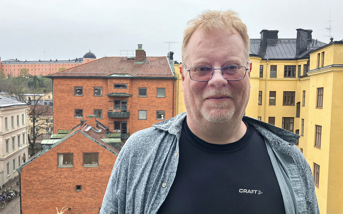 Halvkroppsporträtt på Martin Hansson i skjorta över svart tshirt och krotklippt hår. I Bakgrunden på avstånd hustak och -fasader i tegel och gul puts.