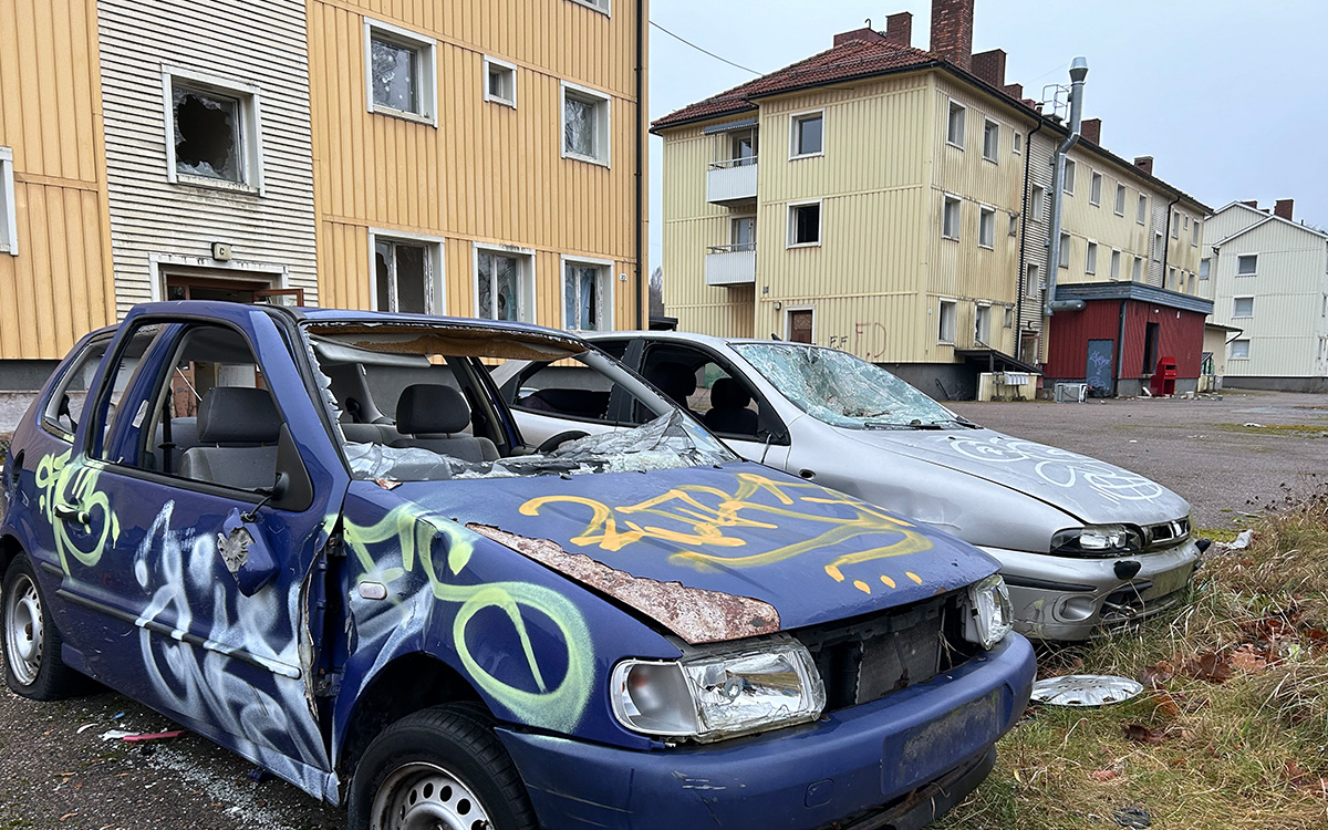 Två skrotbilar i förgrunden. Bakom syns nedgångna hyreshus.