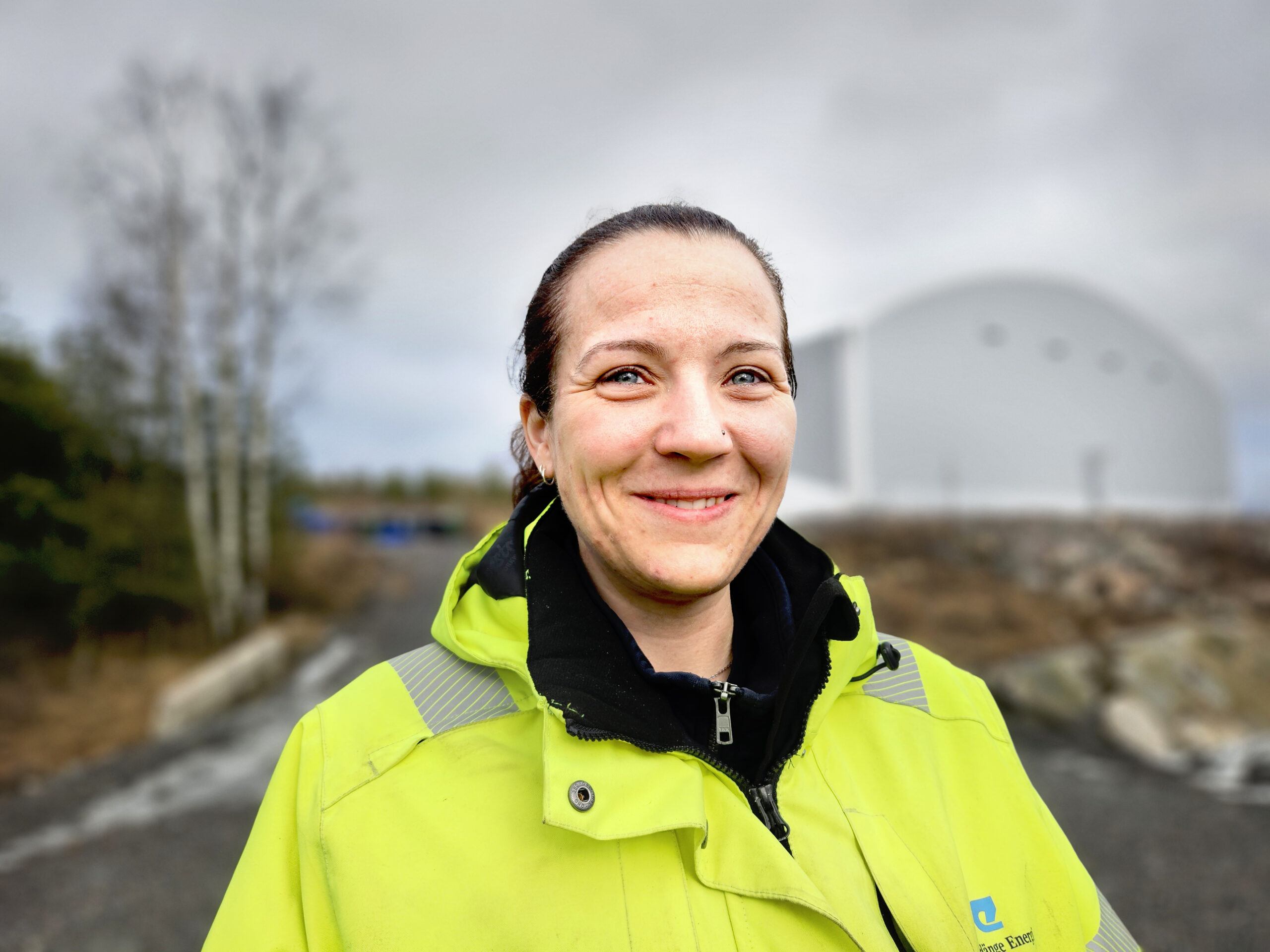 En kvinna i gult arbetsställ med grå himmel och en grå byggnad i bakgrunden.