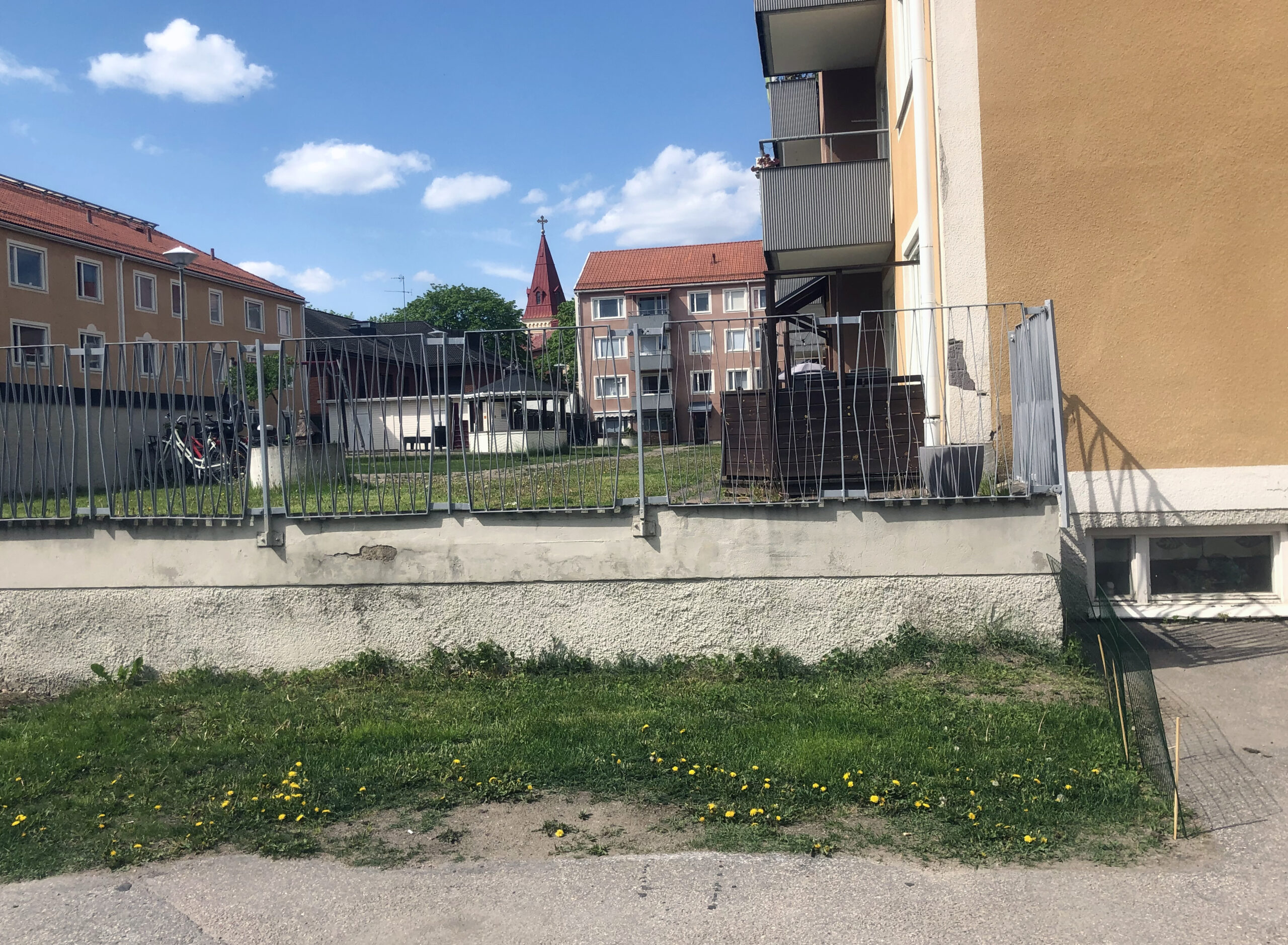 En gräsremsa längs en betongmur med ett staket ovanpå. I bakgrunden hyreshus och ett kyrktorn.