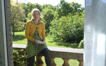 Anna Bengtsson i ljust uppsatt hår, en grön mönstrad klänning med bin på och en gul kofta, halvsitter på ett balkongräcke i sten. I bakgrunden syns en stor gräsmatta och buskar, träd, skog. Anna Bengtsson har forskat kring betydelsen av kontakt med utemiljön. Att ha en uteplats är betydelsefullt på flera sätt för den fysiska och psykiska hälsan och för att känna trygghet.