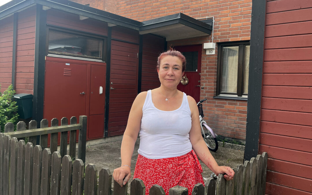 En kvinna i vitt linne och röd kjol står vid ett staket utanför ett radhus i rött tegel. Hennes i grunden mörka hår är färgat rött.