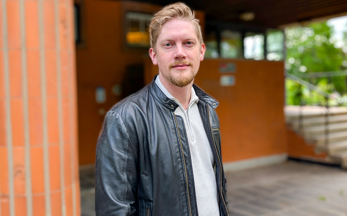 Man som tittar in i kameran. Ses i halvfigur. Han står framför en byggnad men bakgrunden är mycket oskarp.