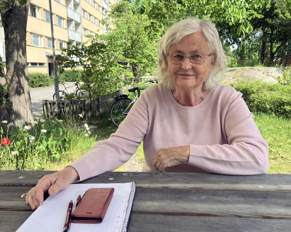 En äldre kvinna med gråvitt hår och glasögon sitter vid ett bord på en uteplats. I bakgrunden ett hyreshus och grönska. Bredvid sig har hon ett block och en mobiltelefon. Hon bär en ljusrosa tröja.