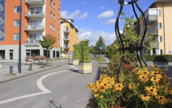 Gatuvy över centrala Knivsta. Blomsterplantering i kruka i förgrunden. Ett orange och ett gult flerbostadshus i bakgrunden.