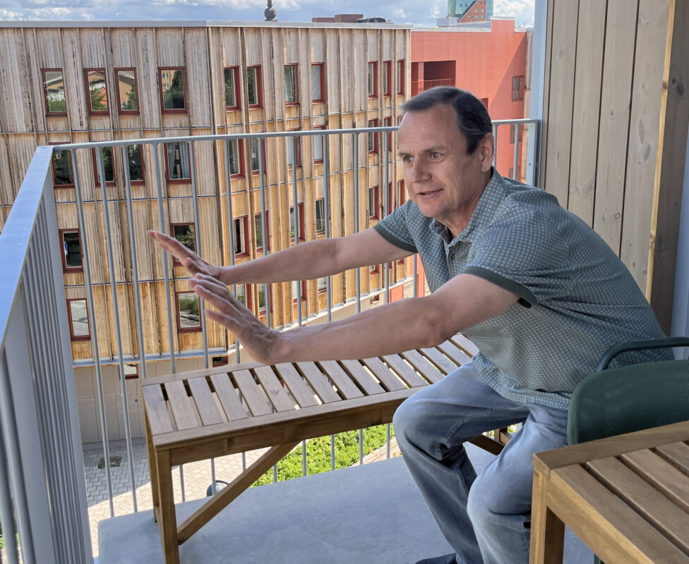 En man i 60-årsåldern står på en balkong. Han böjer på knäna och sträcker händerna framför sig i en yogaövning. Bortanför balkongräcket syns ett nybyggt hus med träfasad.