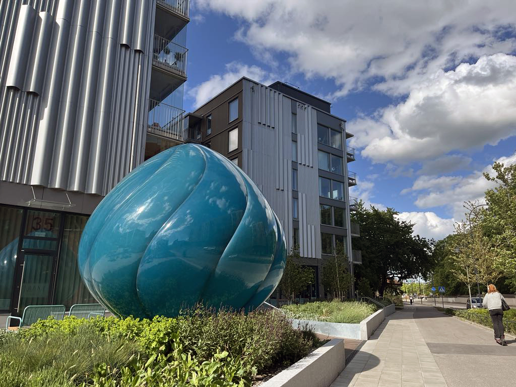 Två nybyggda femvåningshus bredvid varandra. I förgrunden en turkosblå lökformad skulptur.